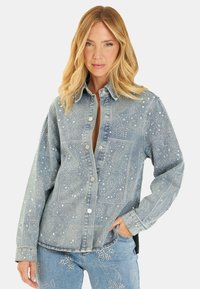 Veste en denim bleu clair avec une fermeture à boutons. Présente des embellissements en argent avec des motifs paisley et étoiles. Manches longues et coupe décontractée.