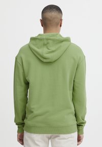 Felpa verde con una grande tasca posteriore e cappuccio con cordini, realizzata in tessuto morbido. Presenta polsini e orlo a coste. Semplice, senza motivi.
