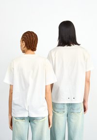 Camisetas de algodón blancas con mangas cortas, que presentan una apertura en la parte posterior y dos botones negros visibles, combinadas con jeans desgastados de color azul claro.