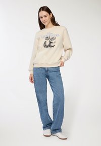 Felpa color crema con grafica di due gattini e la scritta "Coordinatori del Caos", abbinata a jeans dritti blu e sneakers bianche.