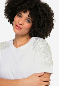 Weißes T-Shirt mit gestickten Blumenmotiven auf den Schultern, verziert mit Pailletten für eine interessante Textur. Rundhalsausschnitt und kurze Ärmel.