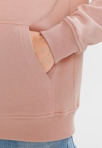Lys rosa hettegenser med kengurulomme og ribbede mansjetter. Teksturen virker myk og jevn, perfekt for hverdagsbruk.
