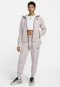 Lichtgrijze Nike sportoutfit met een zip-hoodie en bijpassende joggers. Opvallende zwarte accenten en een cropped witte top eronder.