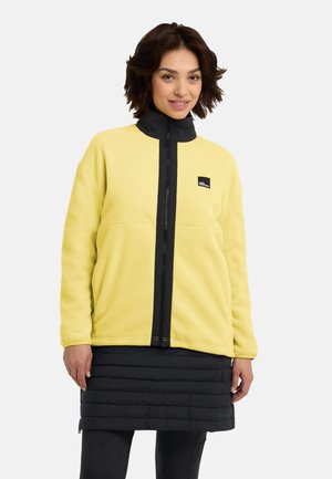 Geel fleece jack met een zwarte ritssluiting en staande kraag, met een logo op de borst en een relaxte pasvorm, gedragen over een zwarte puffy rok.