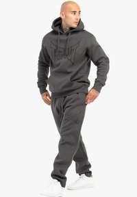 Grå hættetrøje og joggingbuksesæt med et stort, præget "TAPOUT" logo. Stoffet ser blødt ud; har en hætte med snor og sidelommer.