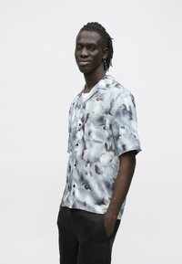 Camisa de manga curta com botão à frente, em azul claro com padrão semelhante a nuvens em tons mais escuros; apresenta um bolso no peito e um ajuste descontraído.