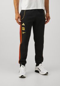 Black athletic joggers with orange and red side stripes, adidas logo, gold accents, and elastic cuffs. Pairing with white sneakers.