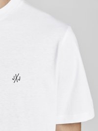 Camiseta de algodón blanca con cuello redondo y mangas cortas. Presenta un pequeño logotipo negro en la esquina superior izquierda. Textura suave.