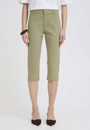 Vrouw die olijfgroene cropped broek, wit topje, zwart armbandje en donkere puntige hoge hakken draagt, staand tegen een effen achtergrond.