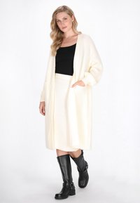 Langer, offener Cardigan in Creme, kombiniert mit einem schwarzen Top und einem cremefarbenen Ripprock. Das Model trägt kniehohe schwarze Stiefel mit Schnallen.
