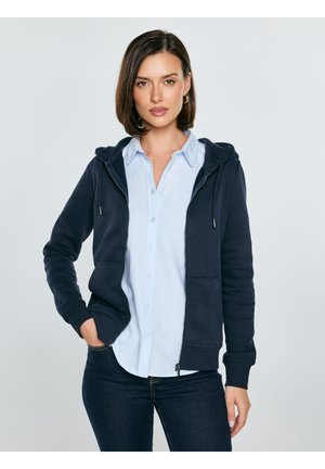 Moteris trumpais rudais plaukais, dėvinti auksinius lankų formos auskarus, šviesiai mėlyną marškinių su sagomis ir tamsiai mėlyną gobtuvą su užtrauktuku, stovinti viena ranka kišenėje.