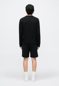 Must pikkade varrukatega särk koos mustade lühikeste pükstega, millel on korduvad tekstimustrid; valged tossud tekstuuriga tallaga ja valged sokid.