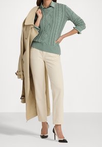 Ljusbeige trenchcoat, texturerad grön kabelstickad tröja, blek skjorta med krage, krämfärgade byxor och högklackade skor i svart och beige.