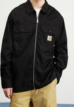 Person iført en sort lynlåstrøje med knaplukning på brystlommerne og et lille Carhartt-logo, kombineret med beige bukser.