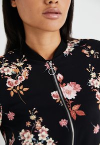 Veste bombers florale avec un fond noir, présentant une fermeture éclair à l'avant et un col côtelé. Le matériau semble lisse ; motifs floraux en rose et jaune.
