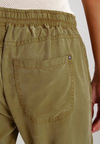 Primer plano de una persona que lleva pantalones verdes oliva con cinturilla elástica y un bolsillo trasero, combinados con una blusa blanca texturizada.