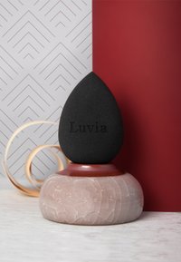 Applicatore a goccia nero su una base testurizzata color rosa, con nome del marchio inciso, posizionato su sfondi decorati.