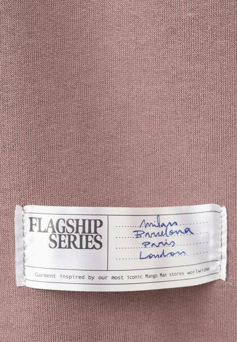 Helles Mauve-Stoff mit einer strukturierten Strickoptik. Das Etikett trägt die Aufschrift "FLAGSHIP SERIES" mit handschriftlichen Städtenamen: Mailand, Barcelona, Paris, London.
