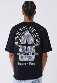 Camiseta de algodón negra con un gran gráfico en la parte posterior con diseño floral, el texto "Alcanza las Estrellas" y "Project X Paris."
