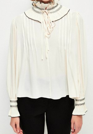 Blouse blanche à manches longues avec des plis, col montant à volants, bordure zigzag noire et lien au col, portée avec un pantalon noir.