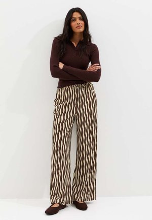 Mujer de pie con los brazos cruzados, vistiendo un suéter acanalado marrón oscuro, pantalones beige de pierna ancha con rayas onduladas marrones y zapatos planos marrones.