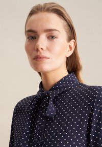 Blusa blu navy con pois bianchi, dotata di colletto, fiocco e chiusura con bottoni sul davanti. Tessuto liscio, vestibilità sartoriale.