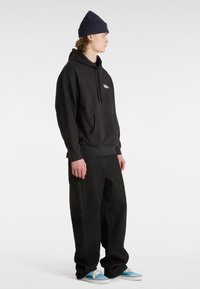 Felpa nera con cappuccio e tasca frontale, con logo bianco, abbinata a pantaloni scuri larghi e sneakers blu, completata da un berretto blu navy.
