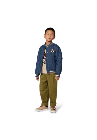 Veste bomber en velours côtelé bleu avec des rayures blanches, écusson sur la poitrine, t-shirt crème avec un graphique de tigre, pantalon vert olive, baskets beiges avec motifs.