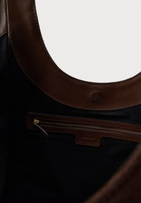 Hnedá kožená taška typu tote s veľkým zaobleným otvorom. Vo vnútri je čierna látková podšívka a vrecko na zips so zlatými kovaniami.