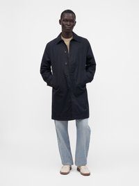 Personne portant un manteau sombre longueur genoux, un jean bleu clair, un pull beige et des baskets blanches, debout les mains dans les poches du manteau.