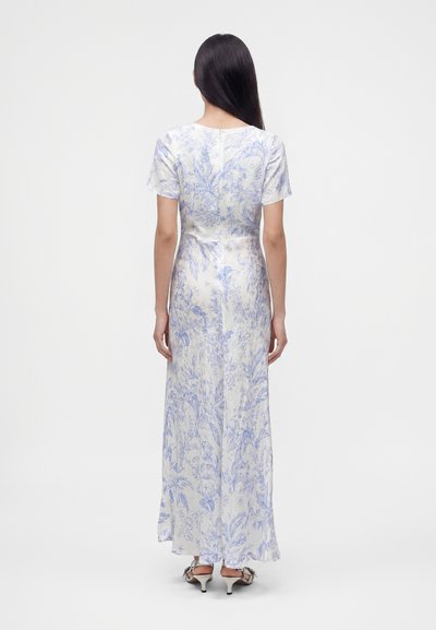 Femme aux longs cheveux foncés portant une robe longue blanche et bleue à fleurs avec manches courtes et des sandales blanches à talons, debout, de dos.