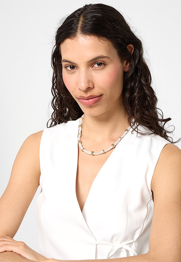 Collana di perline con accenti dorati e elementi bianchi e dorati decorati, indossata da un modello con una maglietta bianca a scollatura a V.