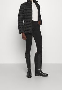 Person som står klädd i en svart puffjacka, vit polotröja, svarta byxor och höga svarta läderstövlar mot en enkel ljus bakgrund.