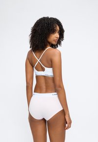 Γυναίκα με σγουρά μαλλιά που φοράει λευκό bralette Calvin Klein με σταυρωτούς ιμάντες στην πλάτη και ταιριαστό λευκό σλιπ, γυρισμένη πλάτη και κοιτάζει πάνω από τον ώμο της.