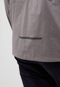 Gray jacket with a textured fabric, featuring a black reflective accent near the hem. Worn over dark pants, showcasing a casual fit.