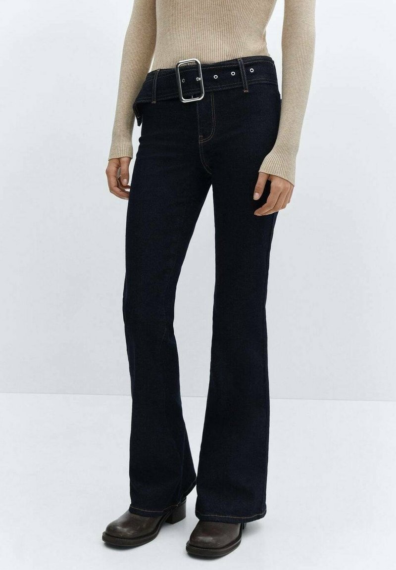 Personne portant un jean évasé foncé avec une large ceinture noire à boucle carrée, un haut côtelé beige à manches longues et des bottes marron foncé.