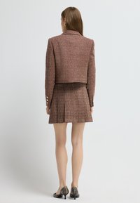 Chaqueta corta de tweed marrón con botones dorados, combinada con una falda plisada a juego. Textura suave, diseño ajustado, hasta la rodilla. Zapatos negros de tacón.