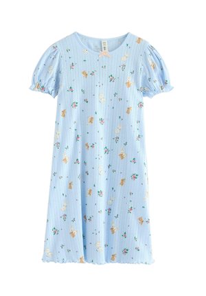 Ung pige stående barfodet iført en lyseblå, kortærmet kjole med blomster- og kaninprint på en hvid baggrund.