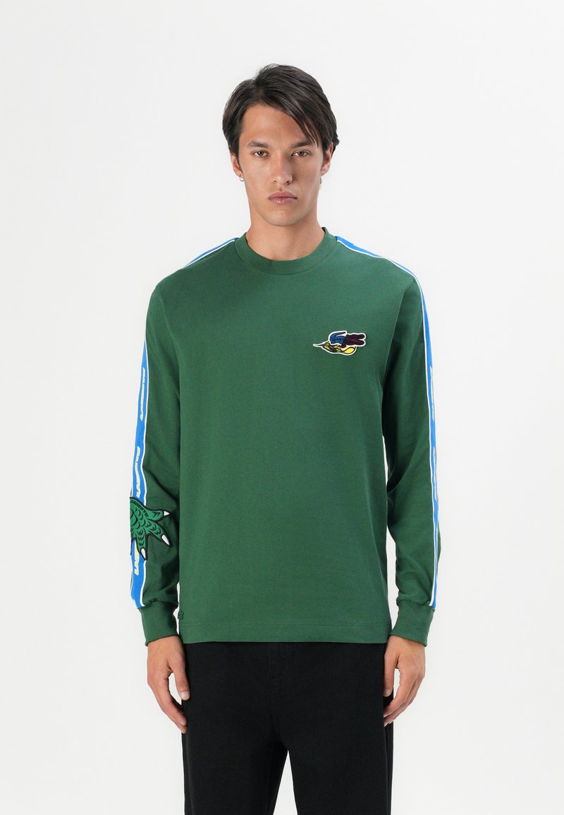 Zielona koszula z długim rękawem z niebieskimi bocznymi paskami, ozdobiona haftowanymi detalami na piersi i rękawach. Wykonana z miękkiego, oddychającego materiału.