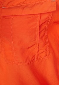 Gros plan sur un tissu orange présentant une poche cousue, une surface texturée et des coutures visibles. Le tissu semble léger et légèrement froissé.