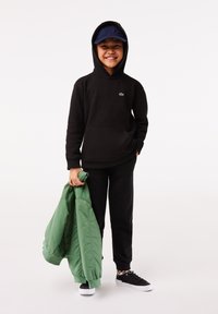 Felpa nera con tasca a marsupio, logo verde Lacoste, pantaloni neri, giacca bomber verde e sneakers nere con suola bianca. Cappellino blu.