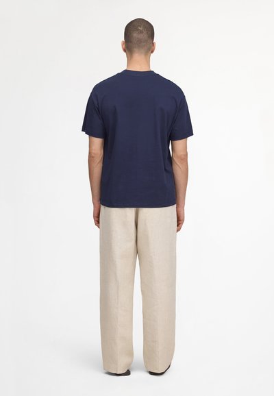 Man standing facing away wearing navy blue short-sleeve t-shirt and loose-fitting beige pants on white background.