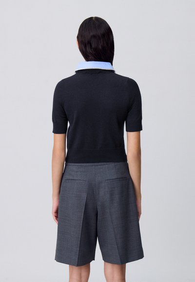 Suéter negro de manga corta con un cuello de camisa azul claro, combinado con pantalones cortos grises a medida con un tejido texturizado y bolsillos laterales.