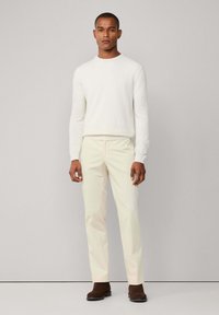 Muškarac u bijelom puloveru s okruglim izrezom, kremastim hlačama i tamnosmešnim cipelama stoji ispred jednolične svijetlosive pozadine.