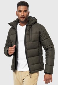 Dunkelgrüne Daunenjacke mit Kapuze, mit gesteppten Abschnitten, schwarzem Reißverschluss und Seitentaschen. Getragen über einem weißen Hemd.