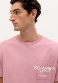 Růžové bavlněné tričko s kulatým výstřihem, krátkými rukávy a náprsní kapsou s bílým nápisem "SCALPERS COMPANY."