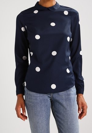 Navy long-sleeve blouse with white polka dots, peter pan collar, worn with light blue jeans.