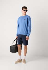 Blue cotton sweatshirt with logo, navy shorts, beige sneakers, and black duffel bag. Model wears black sunglasses, light gray socks visible.