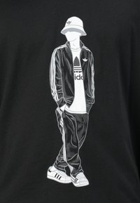 Μαύρη μπλούζα T-shirt με γραφικό ενός ατόμου που φοράει καπέλο bucket, μαύρη φόρμα και λευκά αθλητικά παπούτσια με σχέδιο τριών γραμμών.