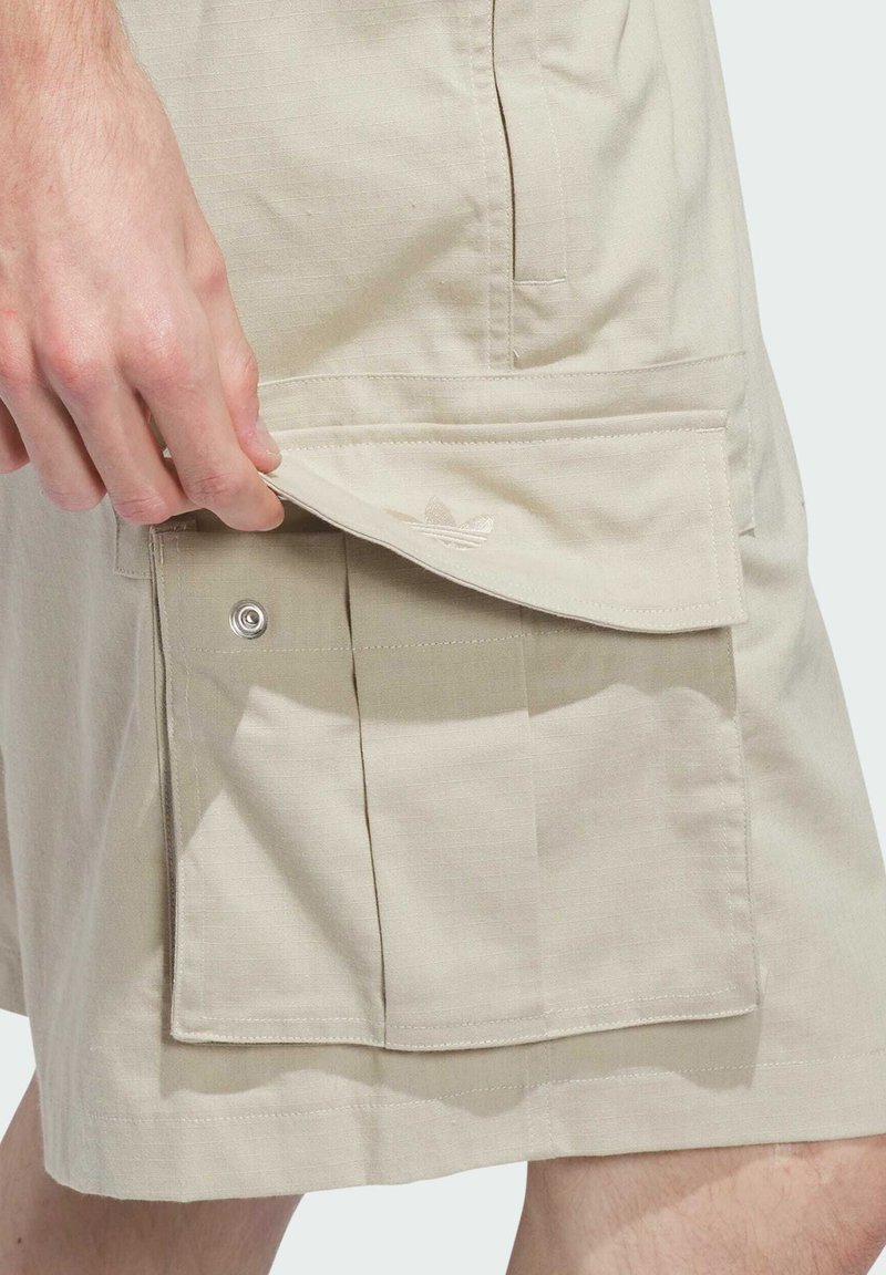 Mano abriendo bolsillo beige de pantalón corto cargo con cierre de botón a presión y logo sutil de Adidas en la solapa del bolsillo.